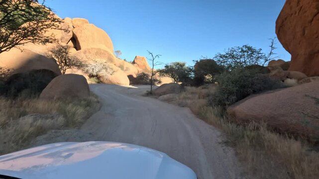 The driver of a lone white car drives along a dusty dirt road among huge boulders in southern Namibia, Africa.