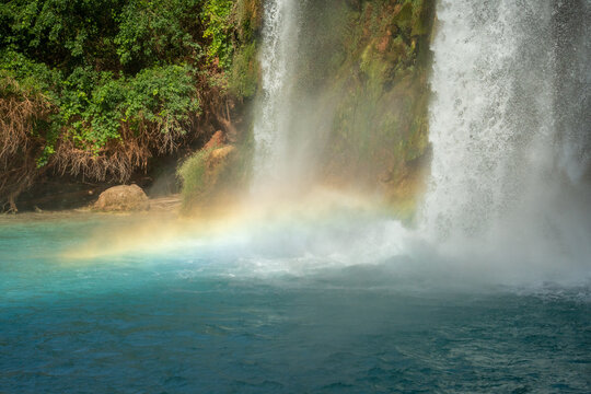 View of Havasu Falls with a rainbow forming in the mist over turquoise water and lush green foliage in Supai, Arizona, United States.
