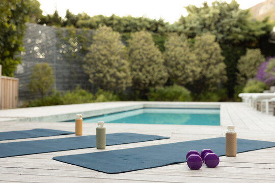 Three dark blue yoga mats are lying on wooden deck by pool, with bottles and dumbbells