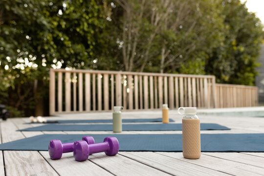 Purple dumbbells are resting on wooden deck by blue mats near pool with bottles, wooden railing