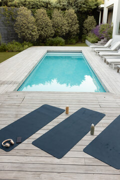 Three dark yoga mats are lying on wooden deck beside lap pool with headphones, phone, bottles