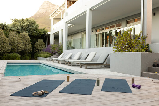 Three dark yoga mats are lying on wooden deck by pool, with bottles, headphones, dumbbell