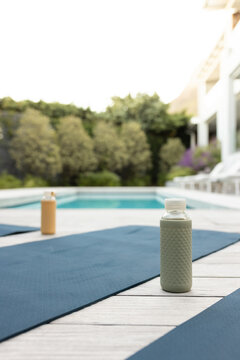 Pale green reusable bottle is standing on wooden deck at poolside with blue mats