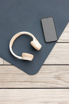 Beige over-ear headphones are resting on dark blue mat angled on wooden deck, black phone nearby