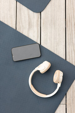 Beige wireless headphones are resting on dark blue yoga mat on wooden deck, black smartphone near