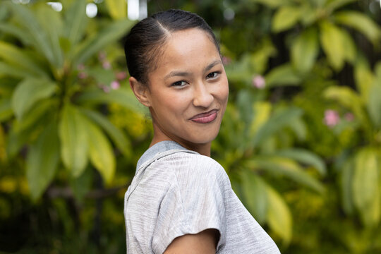Woman wearing gray top, turning over shoulder, looking at camera in garden with pink flowers