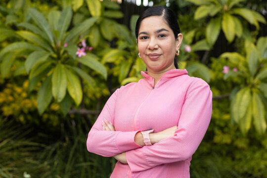 Woman standing with arms crossed in garden among broad leaves, wearing pink zip-up and smartwatch