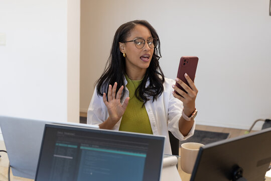 African American woman holding red smartphone, gesturing while speaking at desk with two laptops
