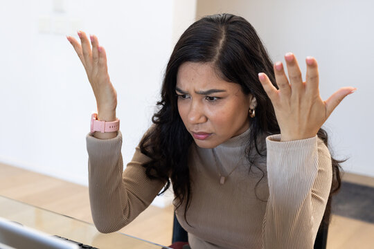 Woman sitting at home office desk, leaning forward, looking at laptop, wearing beige top pink watch