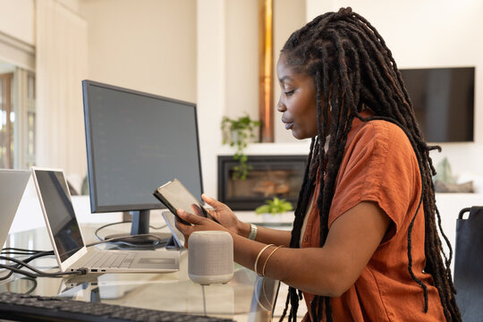 African American woman sitting at glass desk in home office holding phone, using laptop and screen