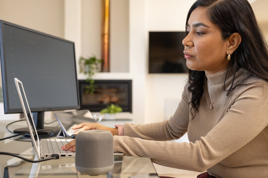 Female in beige turtleneck sitting at glass desk at home workspace, typing on laptop with monitor