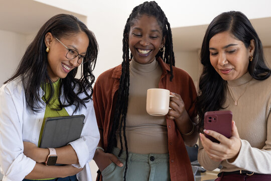 Diverse female coworkers standing in break area holding closed tablet, beige mug and maroon phone