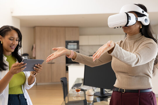 Diverse female pair wearing beige sweater testing VR headset while helper holding tablet in kitchen