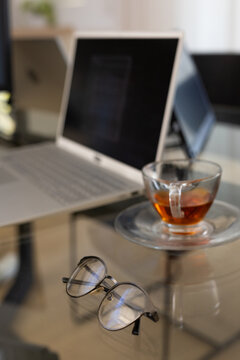 Pair of metal-frame eyeglasses are resting on glass table with teacup and laptop reflections