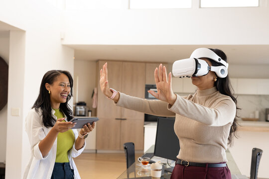 Diverse female friends testing white VR headset and interacting at open kitchen table with tablet