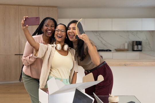 Diverse female friends taking selfie in modern kitchen, holding pink smartphone and clear trophy