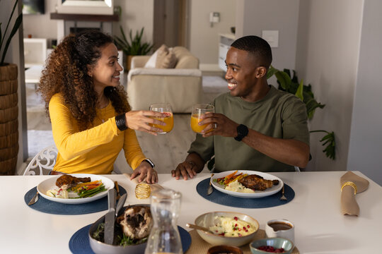 Diverse couple clinking stemless glasses while sitting at home dining area with roasted chicken