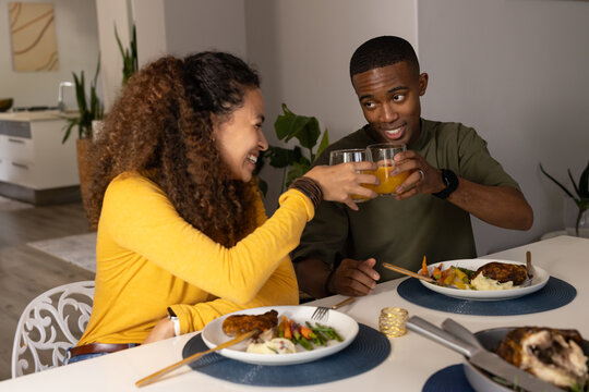 Diverse couple clinking orange juice glasses at home table with plated chicken in olive  and  yellow