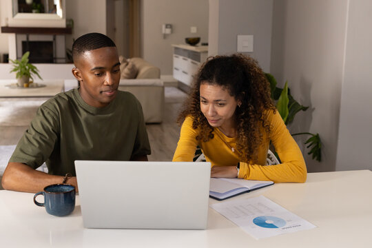 Diverse couple reviewing pie chart on silver laptop at home with notebook and blue mug