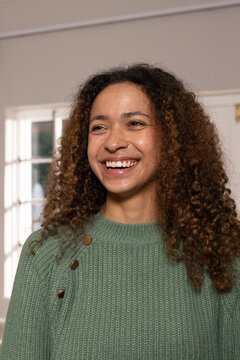 Woman smiling, standing at home by paned window wearing green knit sweater with three metal buttons