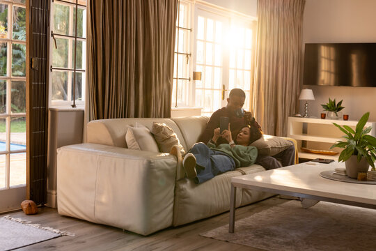 Diverse couple in casual clothes reclining on light leather sofa in living room, sharing smartphone