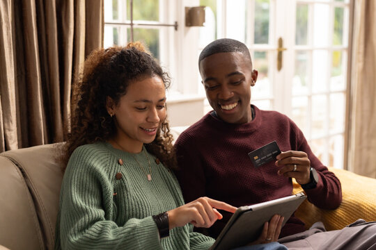 Couple sitting on beige sofa at home, wearing knit sweaters and using tablet with credit card