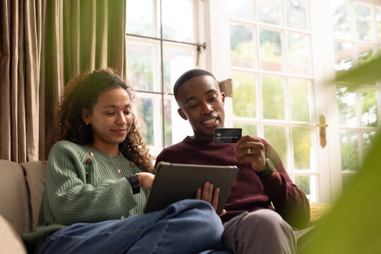 Couple sitting on beige sofa at home sharing tablet, woman in green sweater, man holding card