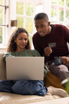 Diverse couple sitting on sofa at home using silver laptop, holding credit card and mug