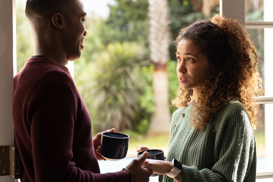 Diverse couple standing in doorway, talking while holding dark mugs, woman wearing smartwatch