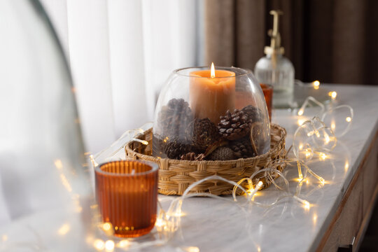 Clear glass cloche is enclosing lit tan pillar candle and pinecones on wicker tray near window