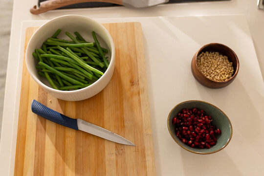 Bamboo board holding white bowl of green stems with blue-knife on white counter under warm light