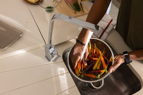 African American man rinsing colorful carrots in mesh colander under chrome faucet at countertop