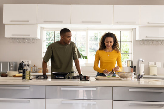 Diverse couple cooking together at home kitchen island with gas cooktop, woman in mustard top