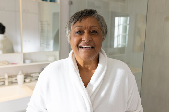 Senior African American woman standing at bathroom vanity smiling pulling white bathrobe near sink