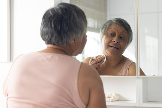 African American senior female wearing pink tank top applying cotton pad to neck at vanity mirror