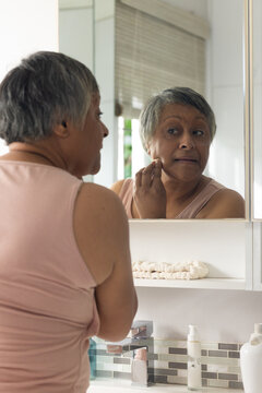 Senior African American woman touching cheek in mirror at bathroom sink wearing pale pink top