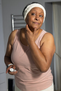 Senior African American woman at bathroom vanity in pink tank applying skincare from white bottle