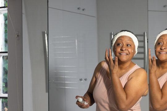 Mature woman at bathroom sink applying cream holding bottle, wearing tank top, headband, copy space