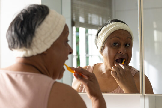 Senior African American woman brushing teeth at vanity mirror with orange toothbrush and headband