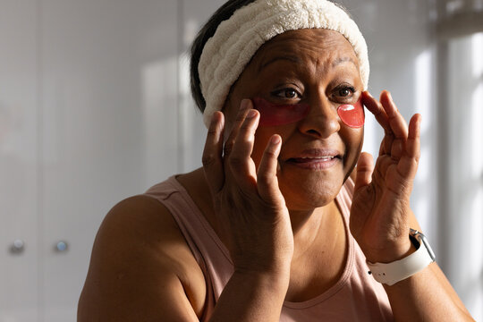 African American woman smoothing red hydrogel under-eye patches at vanity, wearing headband