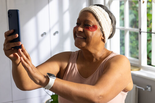 Senior African American woman standing by home window wearing tank, eye patches, holding smartphone