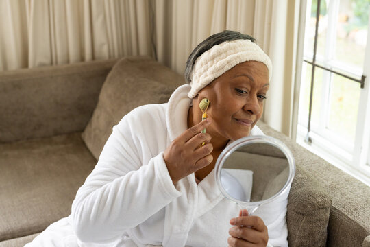 Senior African woman on sofa wearing robe and headband rolling face with gold roller mirror-held