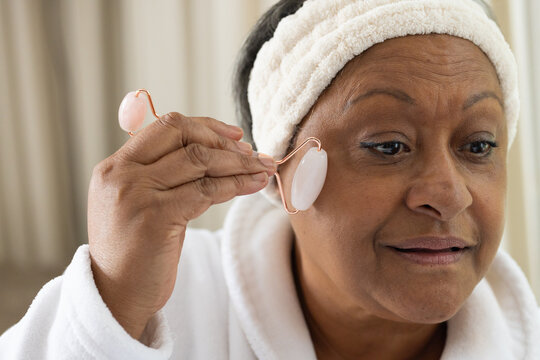 Senior African woman wearing white bathrobe terry headband rolling pink facial roller at vanity