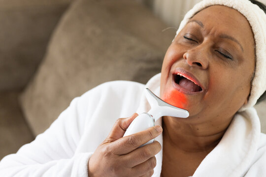 Senior African woman applying LED handheld device to chin on couch in white robe, headband
