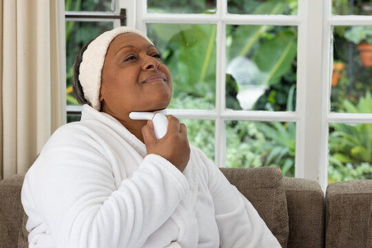Senior African American woman in white bathrobe and terry headband sitting on sofa holding device