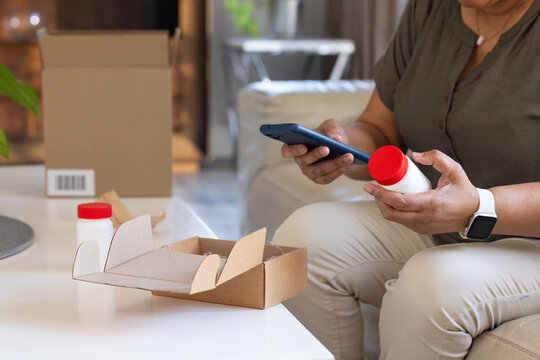 Senior African American woman sitting at home holding bottle and checking phone near cardboard box