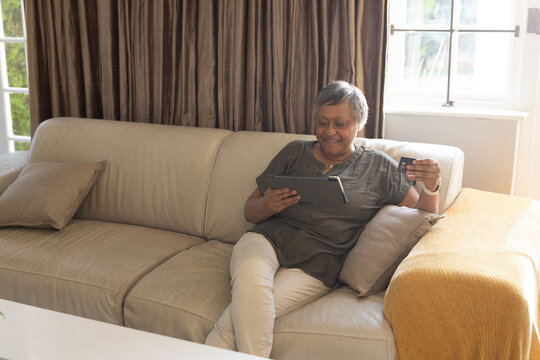 Senior African American woman sitting right on beige sofa checking tablet with card, copy space