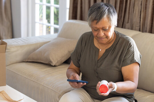 Senior African American woman holding pill bottle and checking phone at home on sofa, copy space