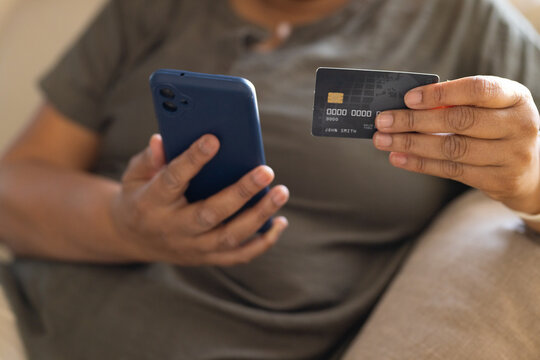 Mature African American woman holding blue-cased phone and tapping bank card chip on sofa at home