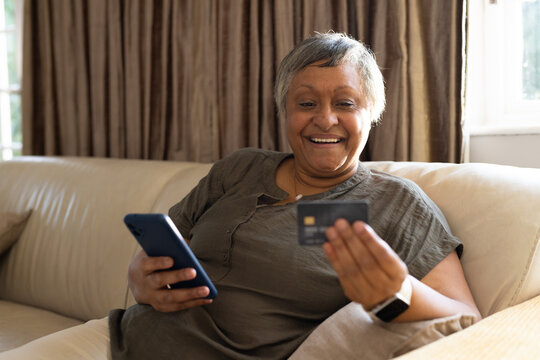 Senior African American woman sitting on beige sofa holding smartphone and payment card near window